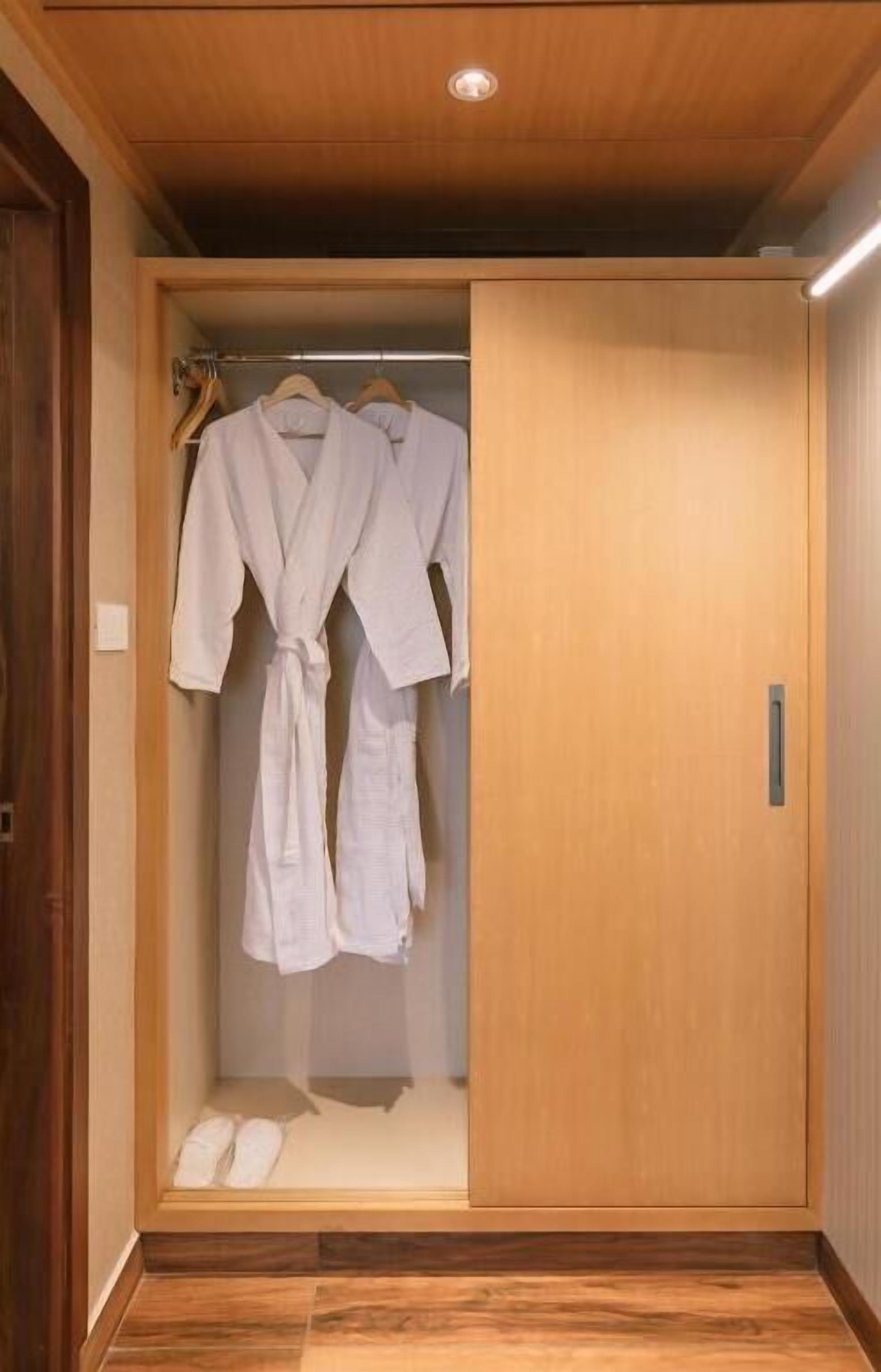 A wooden walk-in closet with a sliding door, two white robes hanging on a rack, and a pair of slippers on the floor.