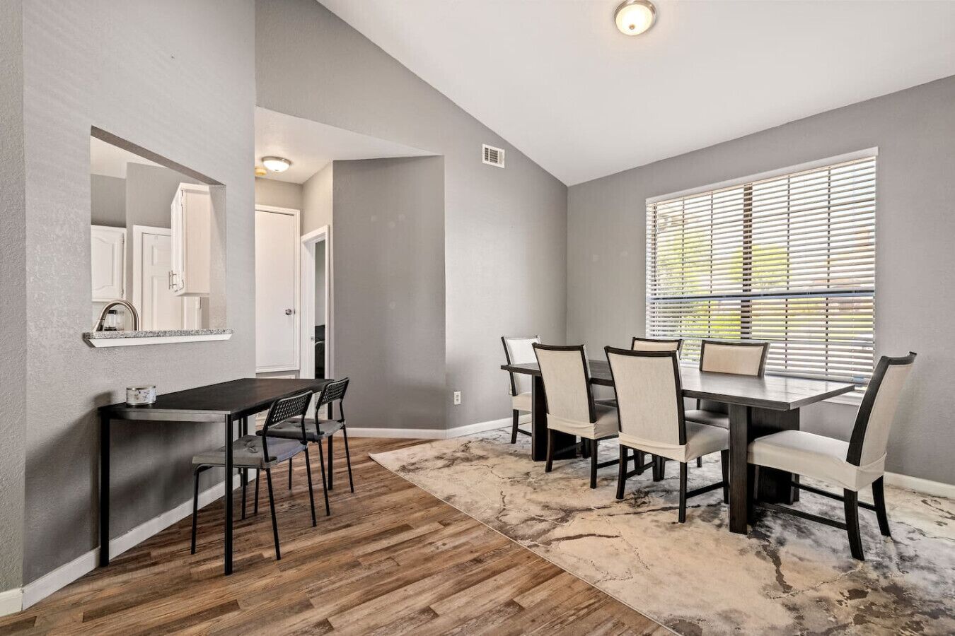 A dining area with a table and chairs, a mirror, a window with blinds, and a wooden floor.