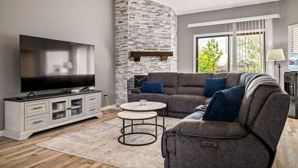 Sala de estar con televisión de pantalla plana, sofá gris, armario blanco, pared de acento de piedra y ventana con persianas.