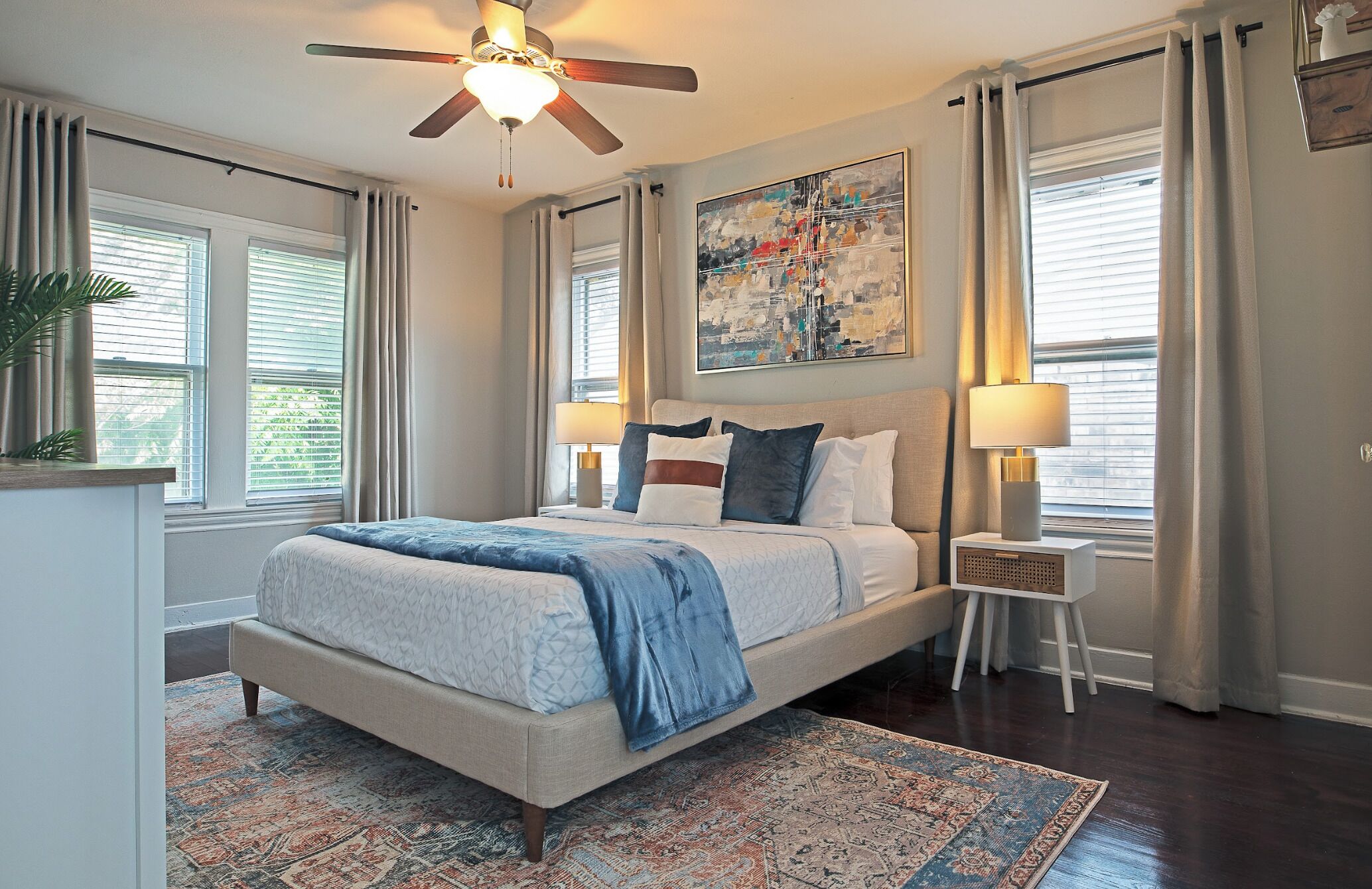 A bedroom with a bed, bedside tables, a ceiling fan, a painting, and a window with curtains.