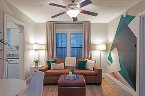 Ein modernes Wohnzimmer mit Deckenventilator, einer Couch mit Kissen, einem Beistelltisch mit Lampe und einem Fenster mit Vorhängen.