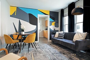 Ein modernes Wohnzimmer mit einem dunklen Sofa, gelben Sesseln, einem Esstisch und einem großen abstrakten Wandbild.
