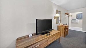 A hotel room with a wooden desk, a flat-screen TV, a mirror, a lamp, a coat rack, and a window with a curtain. - Eagle Motel Suite 105 (Manitou Springs)