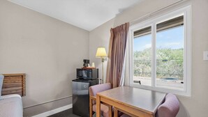 A small room with a bed, a table, a chair, a microwave, a lamp, and a window with curtains. - Eagle Motel Suite 105 (Manitou Springs)