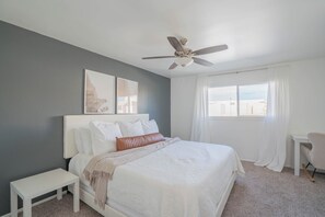 Ein Schlafzimmer mit Bett, Deckenventilator, Fenster mit Vorhängen, Nachttisch und Sessel.