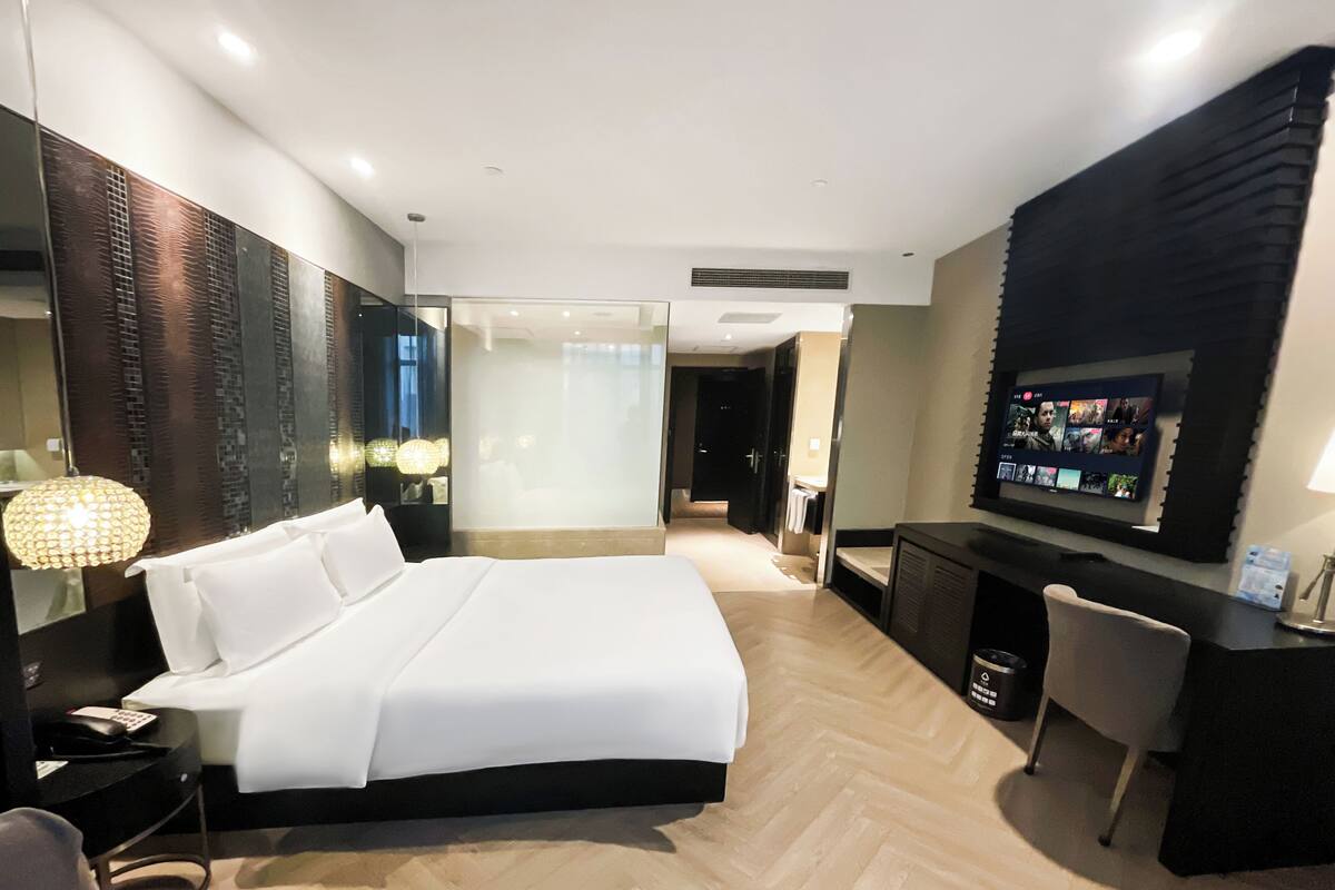 Habitación de hotel moderna con una cama grande, un escritorio con televisor de pantalla plana, una zona de estar separada y un espejo amplio.