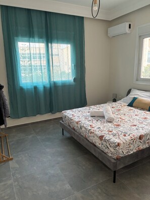 A bedroom with a bed, a window with curtains, an air conditioning unit, and a tiled floor. - Coral Urbanite (Rodos)