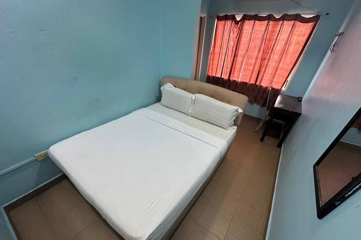 A single bed in a room with light blue walls, a window with red curtains, and a small table with a chair.