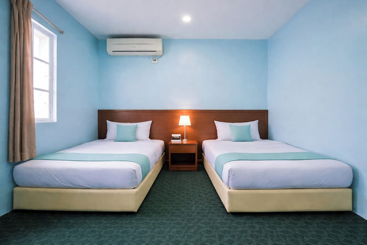 A hotel room with two beds, a nightstand with a lamp, an air conditioning unit, and a window with curtains.