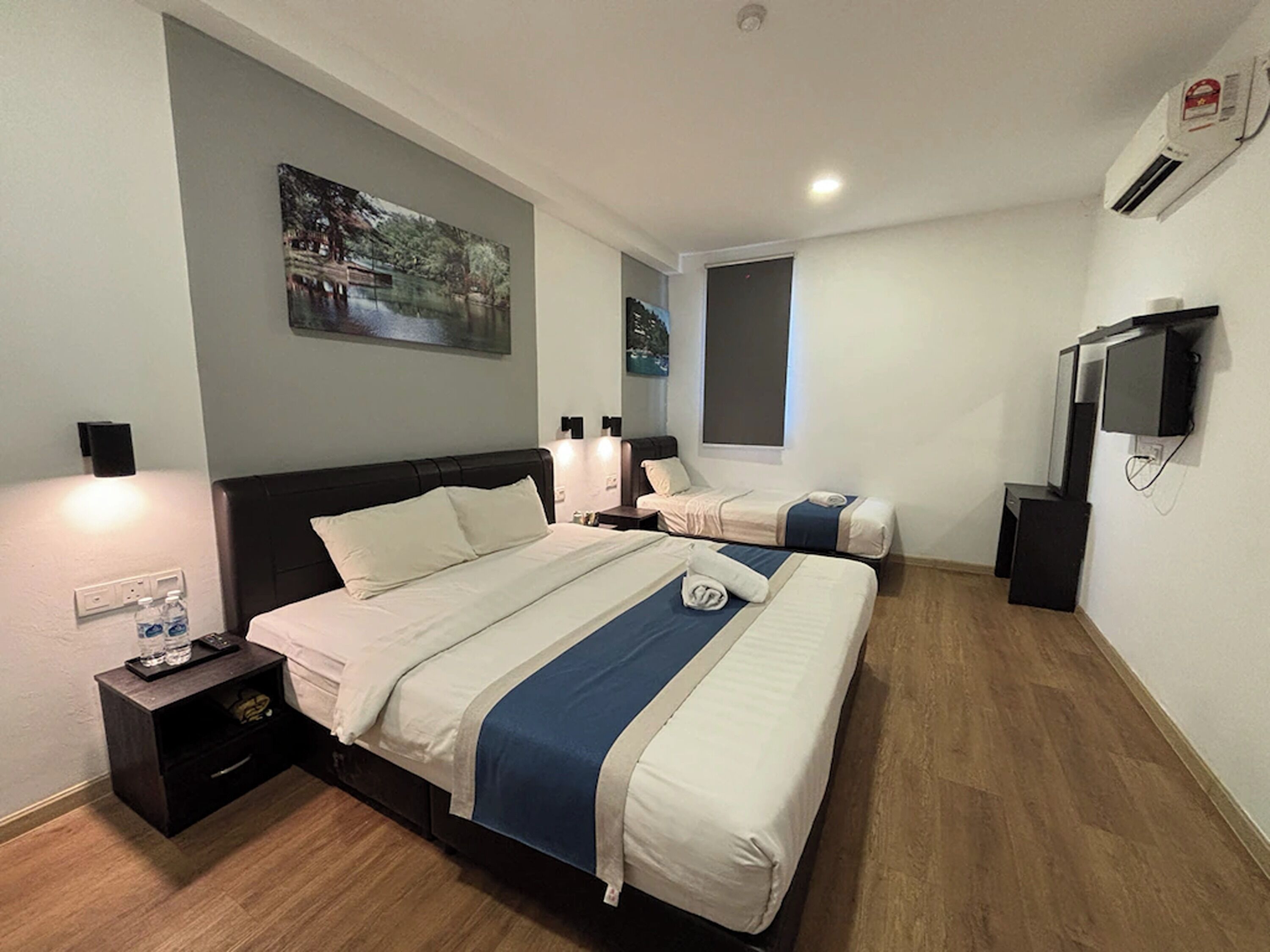 A hotel room with two beds, a nightstand, a wall-mounted TV, and a painting.