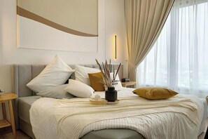 Habitación de hotel ordenada con una cama, almohadas, una mesita de noche, un florero con plantas y una ventana con cortinas.