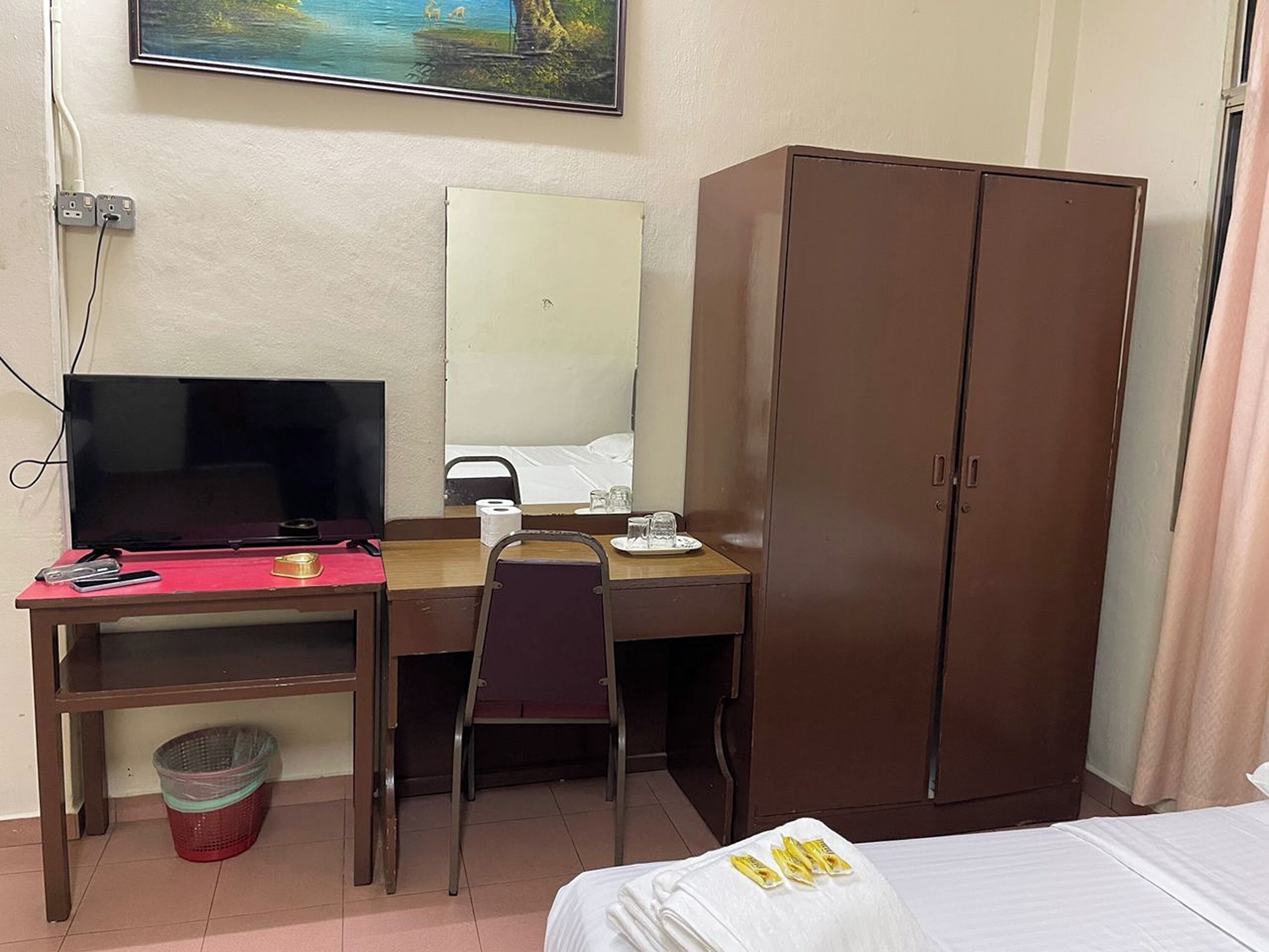 A hotel room with a bed, a desk with a TV, a wardrobe, and a window with a painting.