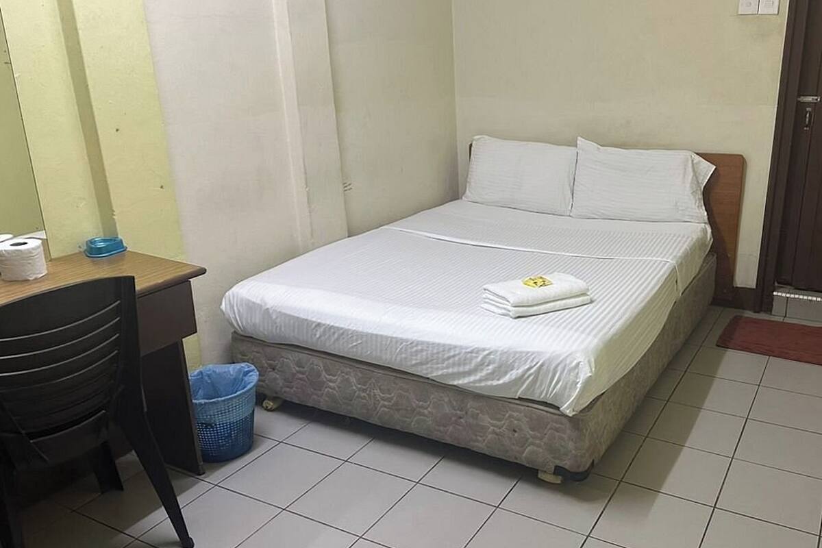 A single bed with white bedding, a wooden headboard, a desk with a chair, and a blue trash bin.