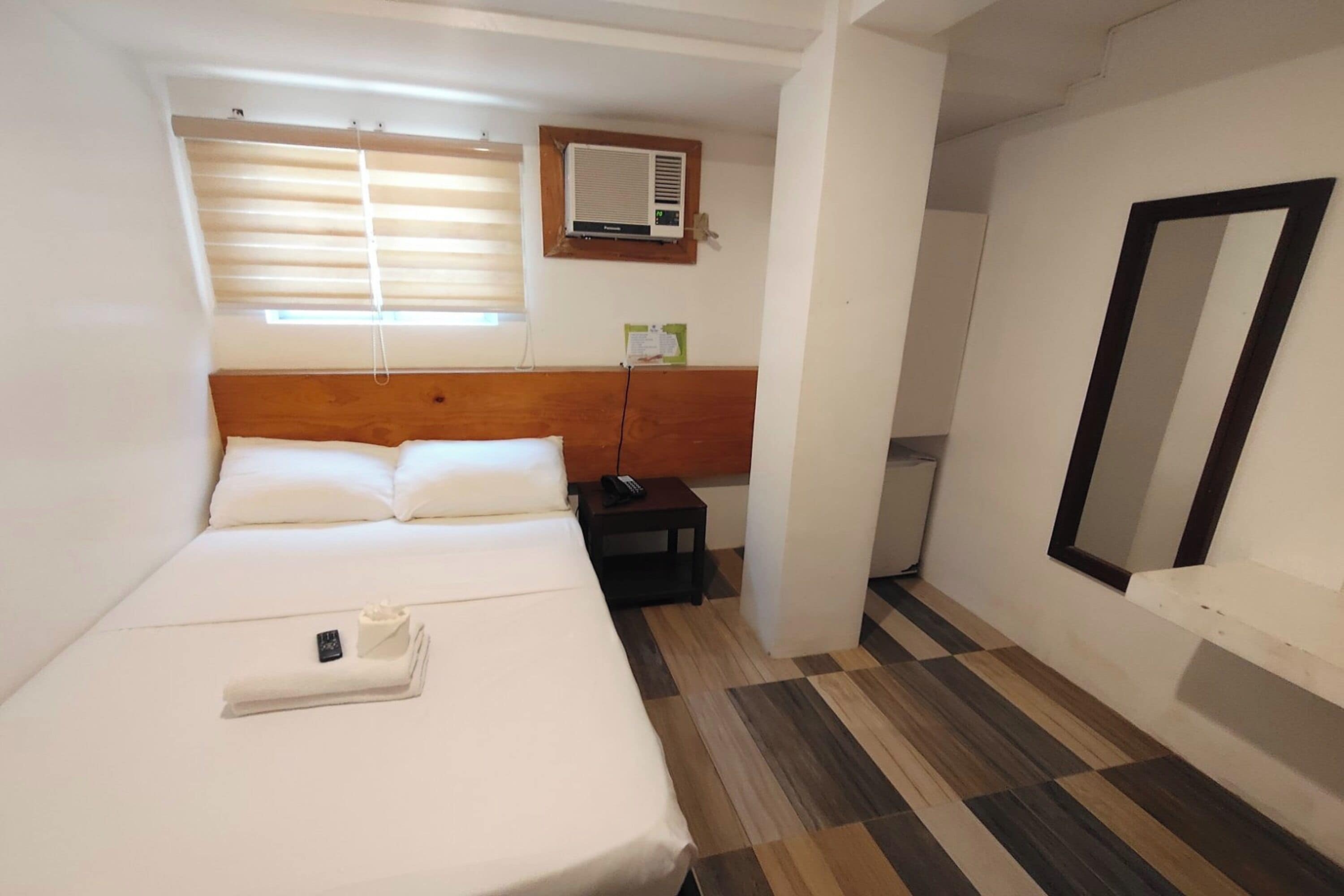 A hotel room with a bed, a bedside table, a mirror, and an air conditioning unit.