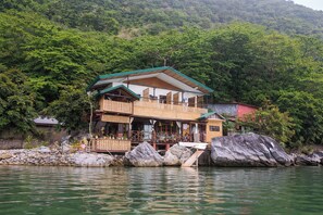 Un edificio de dos pisos con balcón, ubicado junto a una masa de agua con rocas al frente.