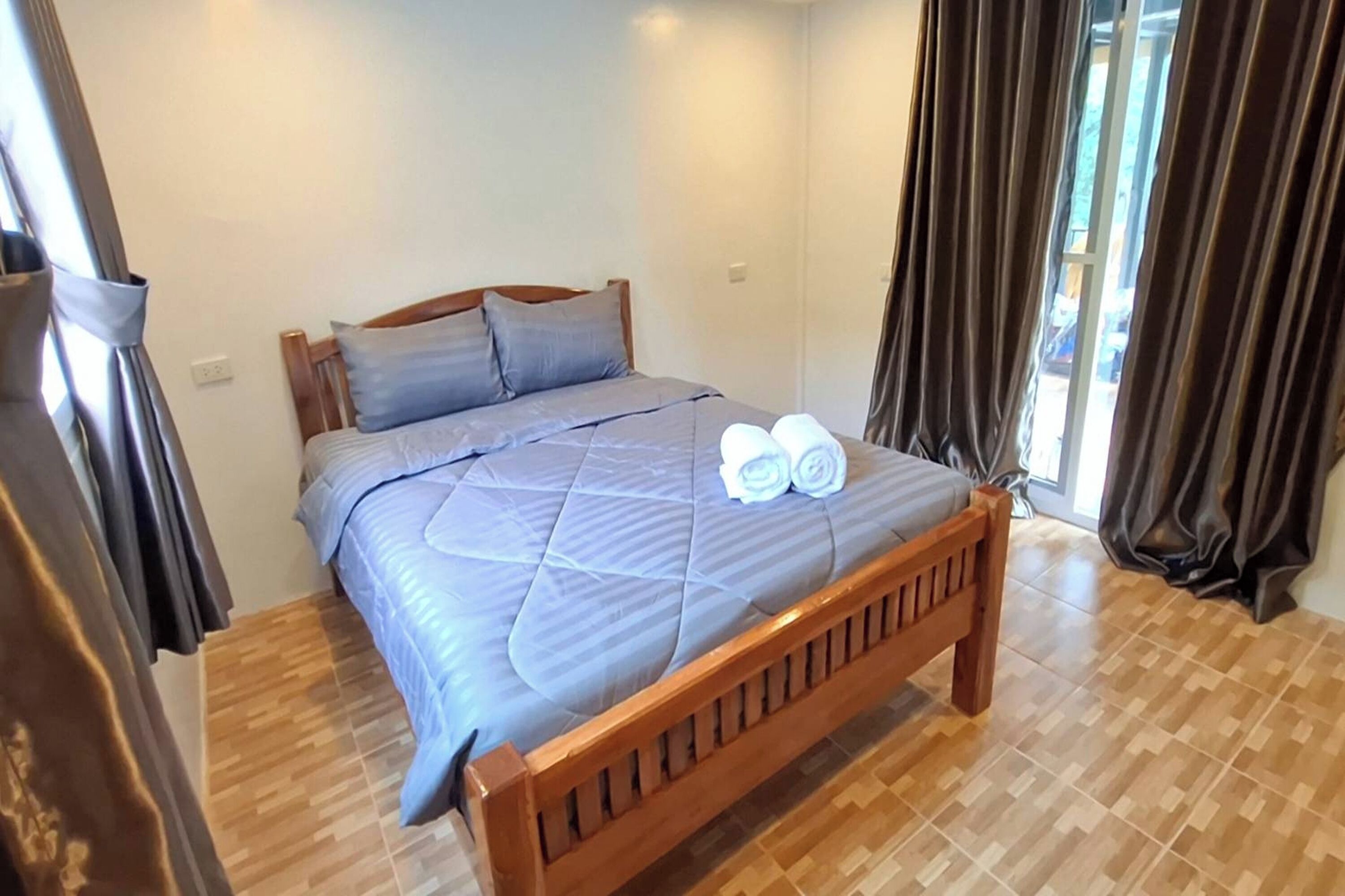 Una cama bien hecha con cubrecolcha acolchado y toallas enrolladas, cabecera de madera y suelo de baldosas.