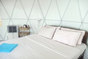 A bed with a white sheet and two pillows, a blue towel, a small table with a kettle, and a wall-mounted fan. - Rai Doi Chang Moncham (Mae Rim)