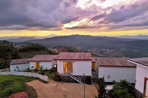 A hillside view of a small cluster of white buildings with a wooden roof, surrounded by greenery and overlooking a valley. - Rai Doi Chang Moncham (Mae Rim)