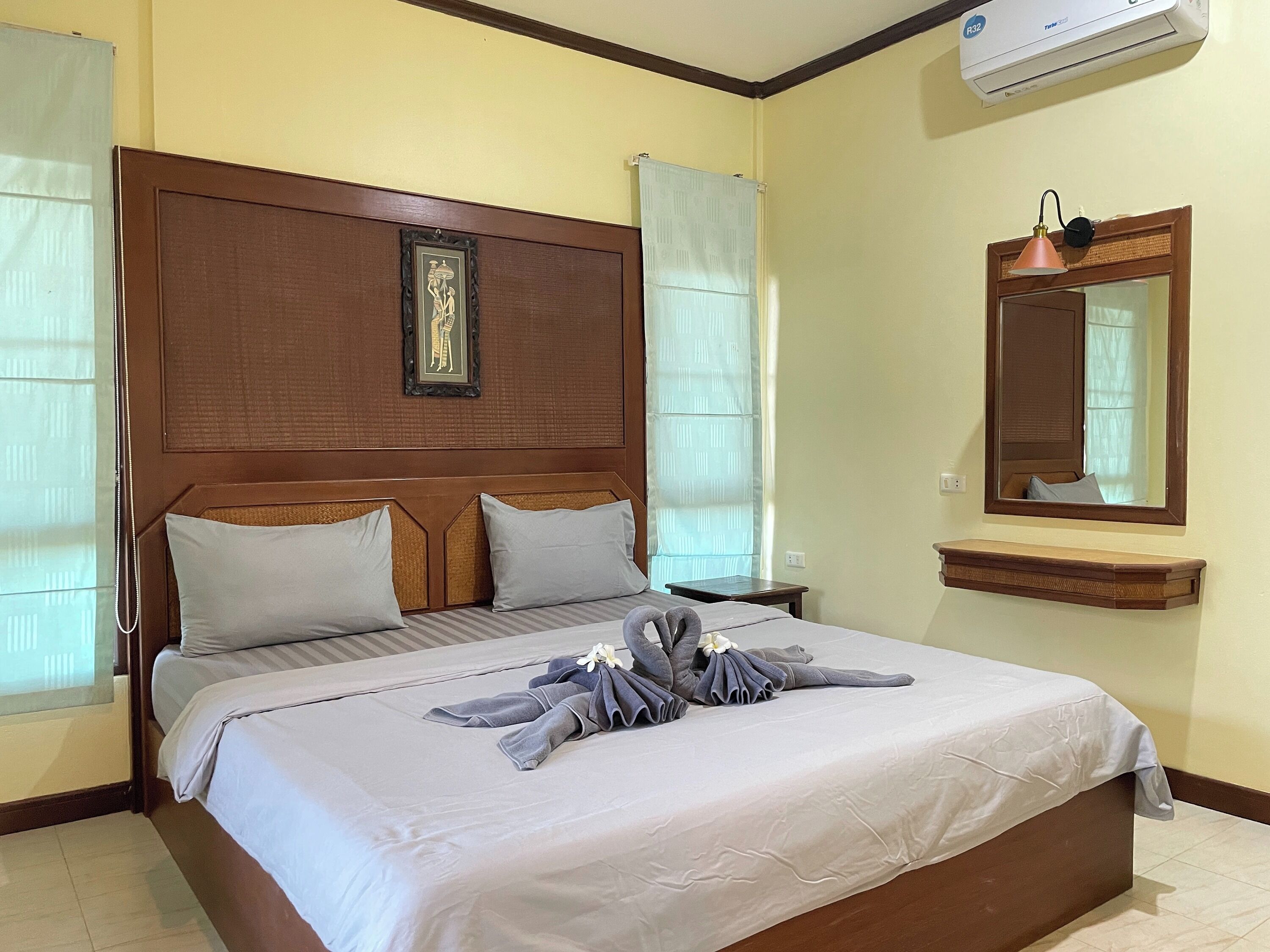 A hotel room with a large bed, a headboard with a framed picture, a bedside table, a mirror, and an air conditioning unit.