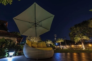 Un espace extérieur aménagé avec des sièges disposés en cercle, protégé par un grand parasol, entouré de verdure et éclairé par un éclairage d’ambiance.