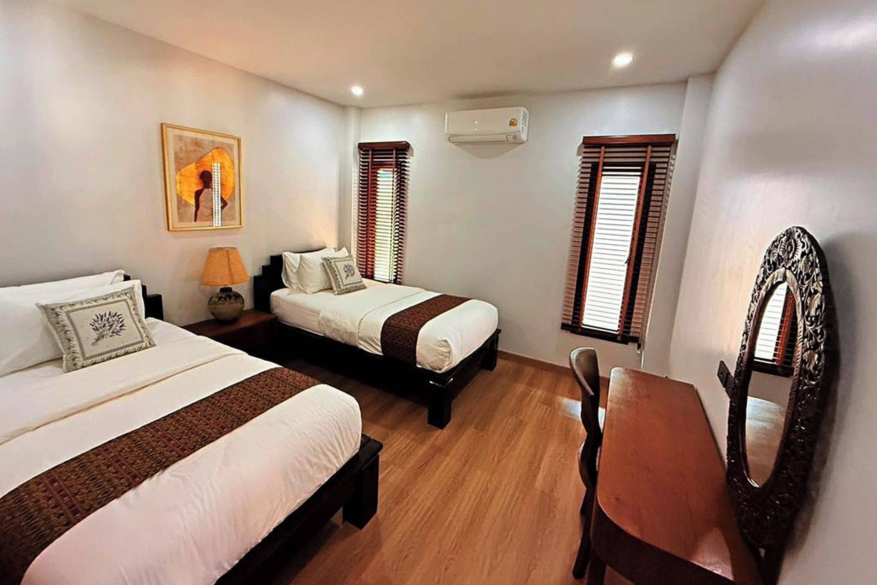 Habitación de hotel con dos camas, piso de madera, escritorio, silla, lámpara, espejo y un cuadro en la pared.