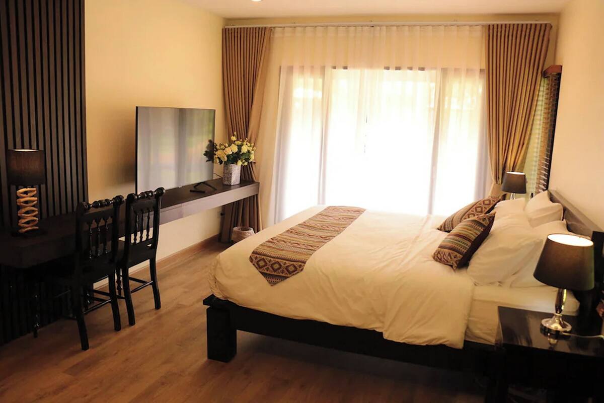 A hotel room with a bed, desk, chairs, a TV, and a window with curtains.