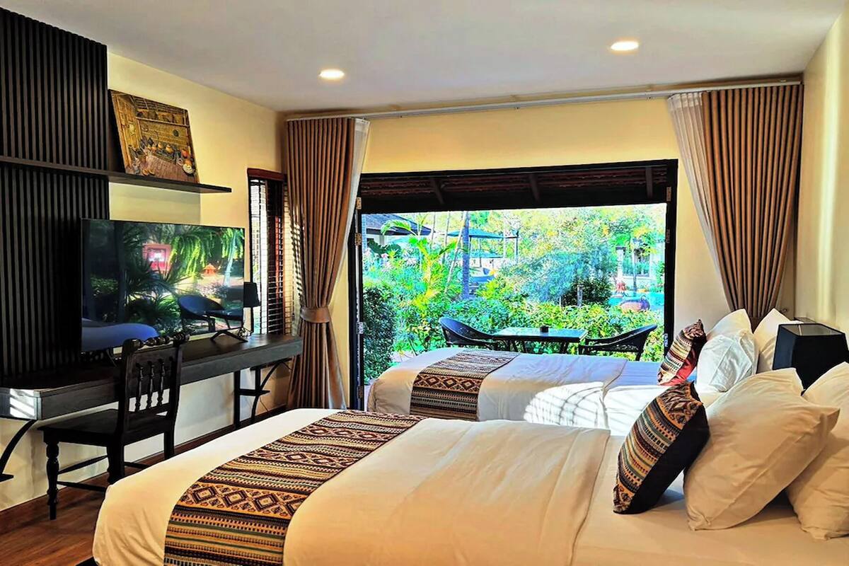 A hotel room with a bed, a desk with a TV, and a view of greenery outside.