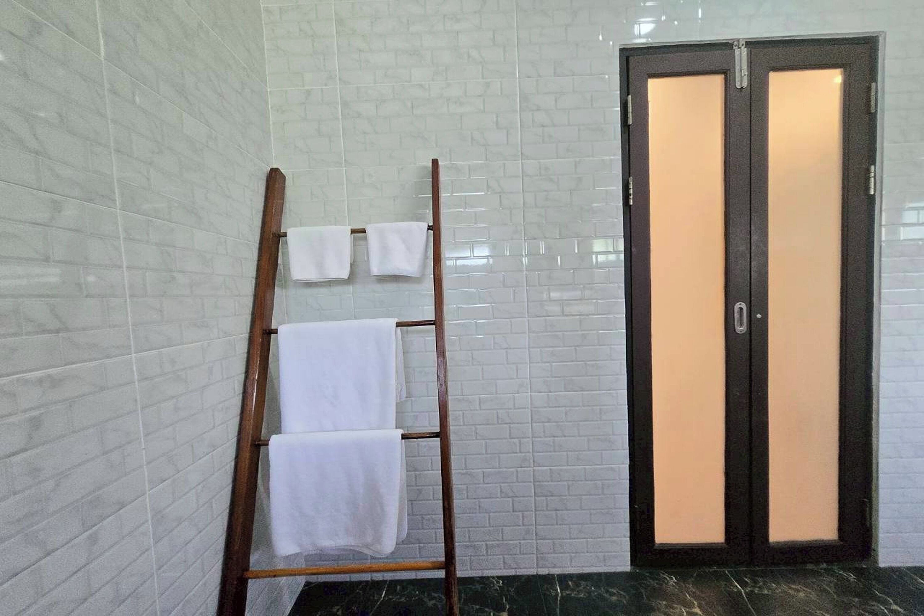 A bathroom with white tiled walls, a wooden towel rack with three towels, and a door with frosted glass panels.