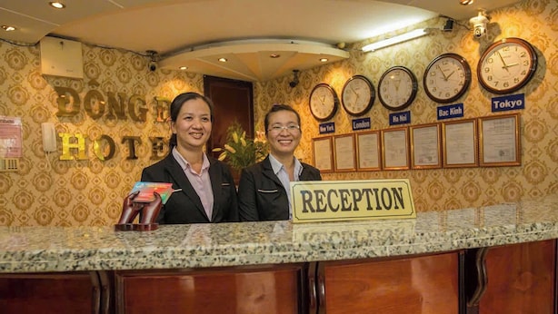 Ein Hotelrezeptionstresen mit einem 'RECEPTION'-Schild, Uhren, die die Zeit anzeigen, und einer Wand mit Städtenamen wie 'London' und 'Tokio'.