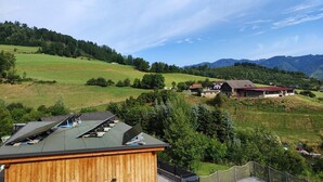 A rural landscape with a wooden building in the foreground, a house with a red roof in the middle ground, and mountains in the background.