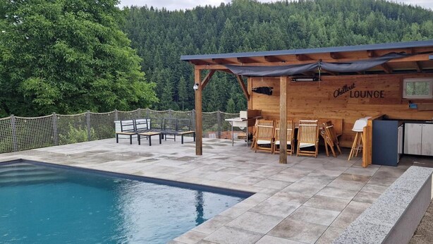 A wooden lounge with a pool, outdoor seating, and a mountainous backdrop.