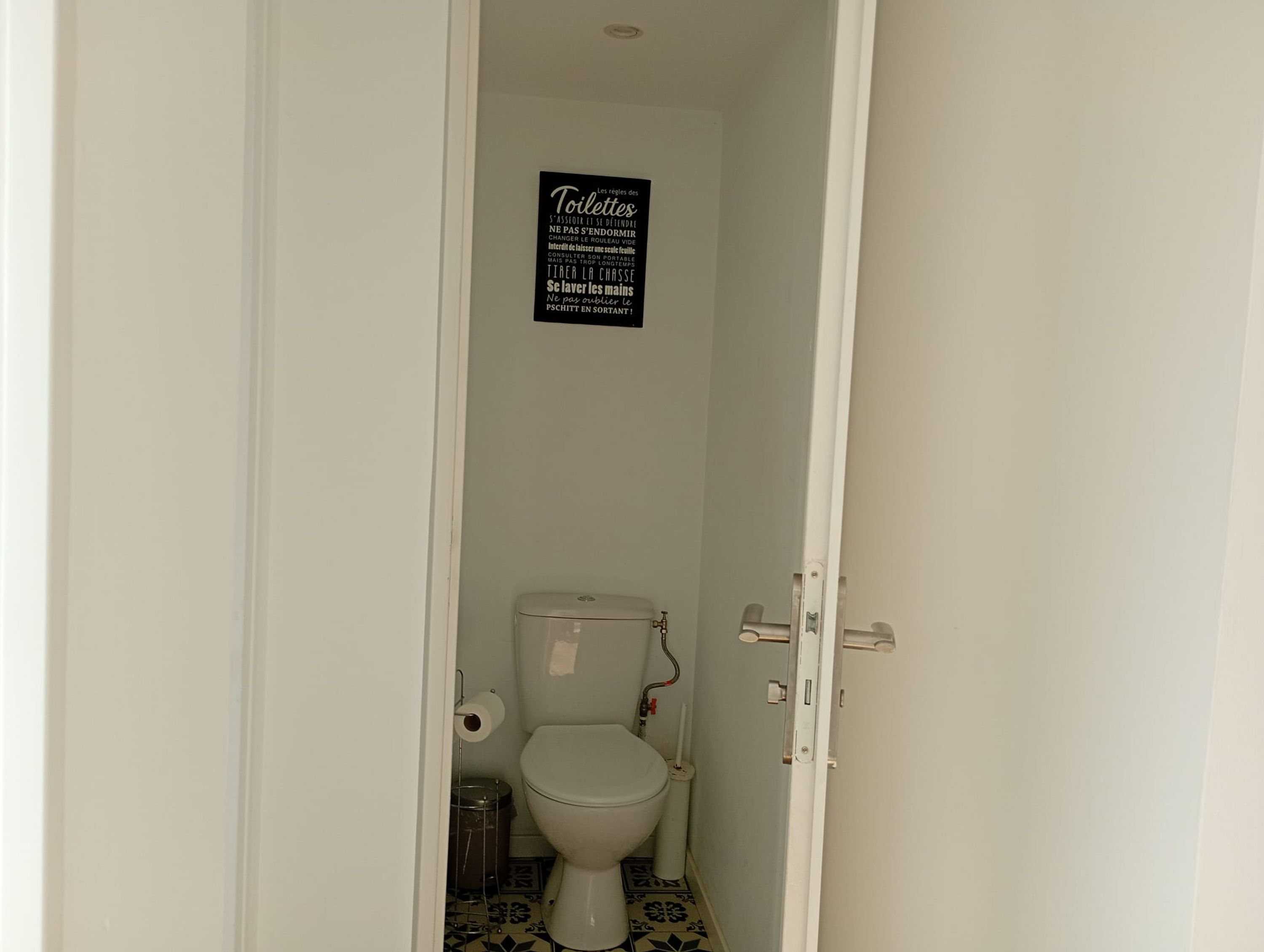 Et lille, aflangt badeværelse med et toilet, toiletpapir og et skilt med fransk tekst.