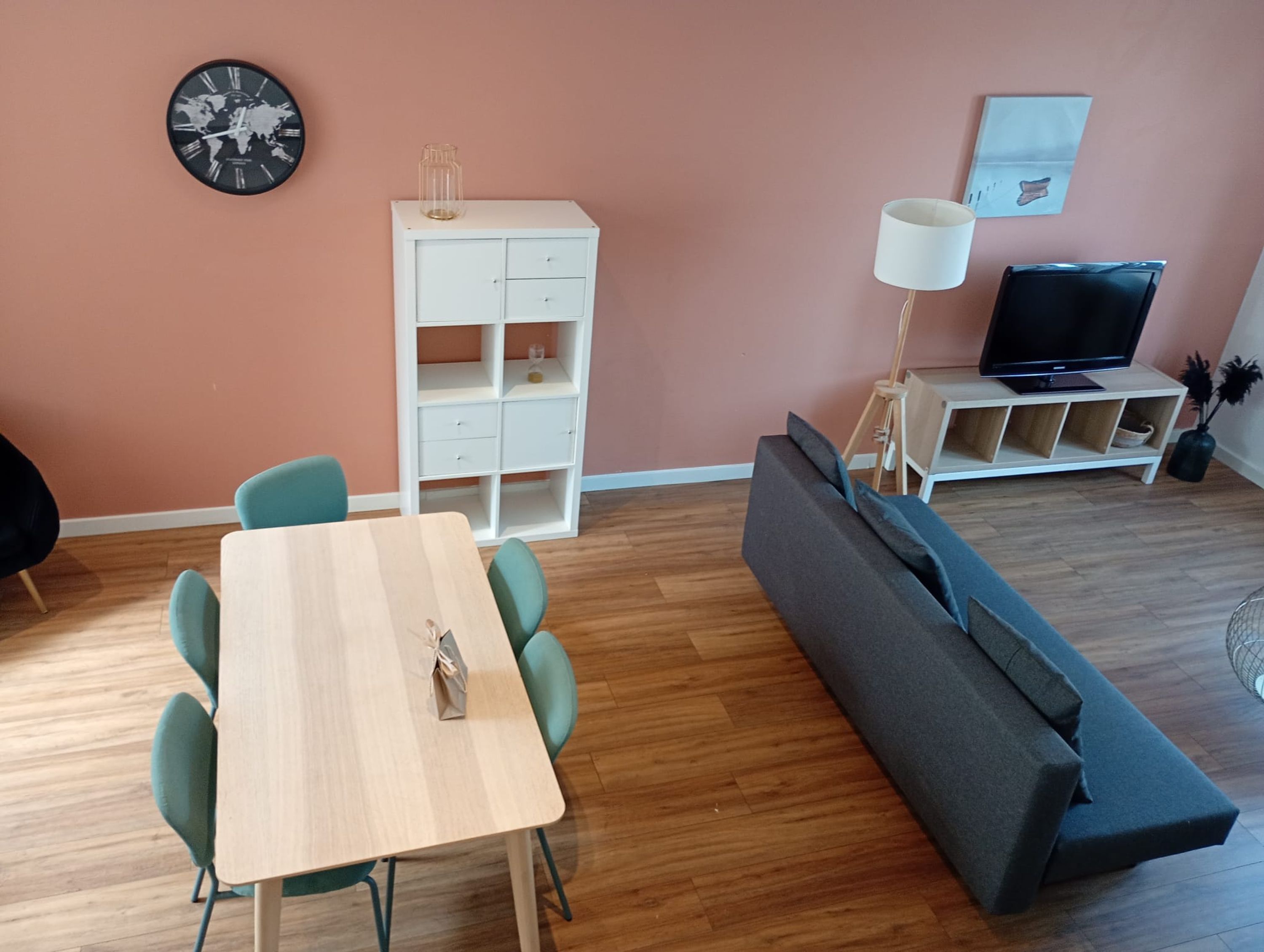 A living room with a wooden table, teal chairs, a grey sofa, a white cabinet, a globe clock, a TV, and a lamp.