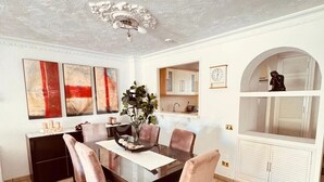 Une salle à manger avec une table en bois foncé, six chaises, un lustre et, au fond, un coin cuisine.