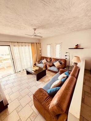 A living room with a brown leather sofa, a glass coffee table, and a ceiling fan. - Spacious 2-bedroom apartment with heated pool, close to the beach (Los Cristianos)