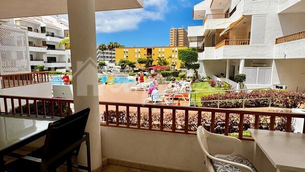 Un balcon donnant sur une piscine, un coin repas avec des chaises et une table, et un bâtiment avec des balcons en arrière-plan.