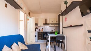 Een ruimtelijk ingerichte leefruimte met kitchenette, eethoek en een bank met decoratieve kussens.