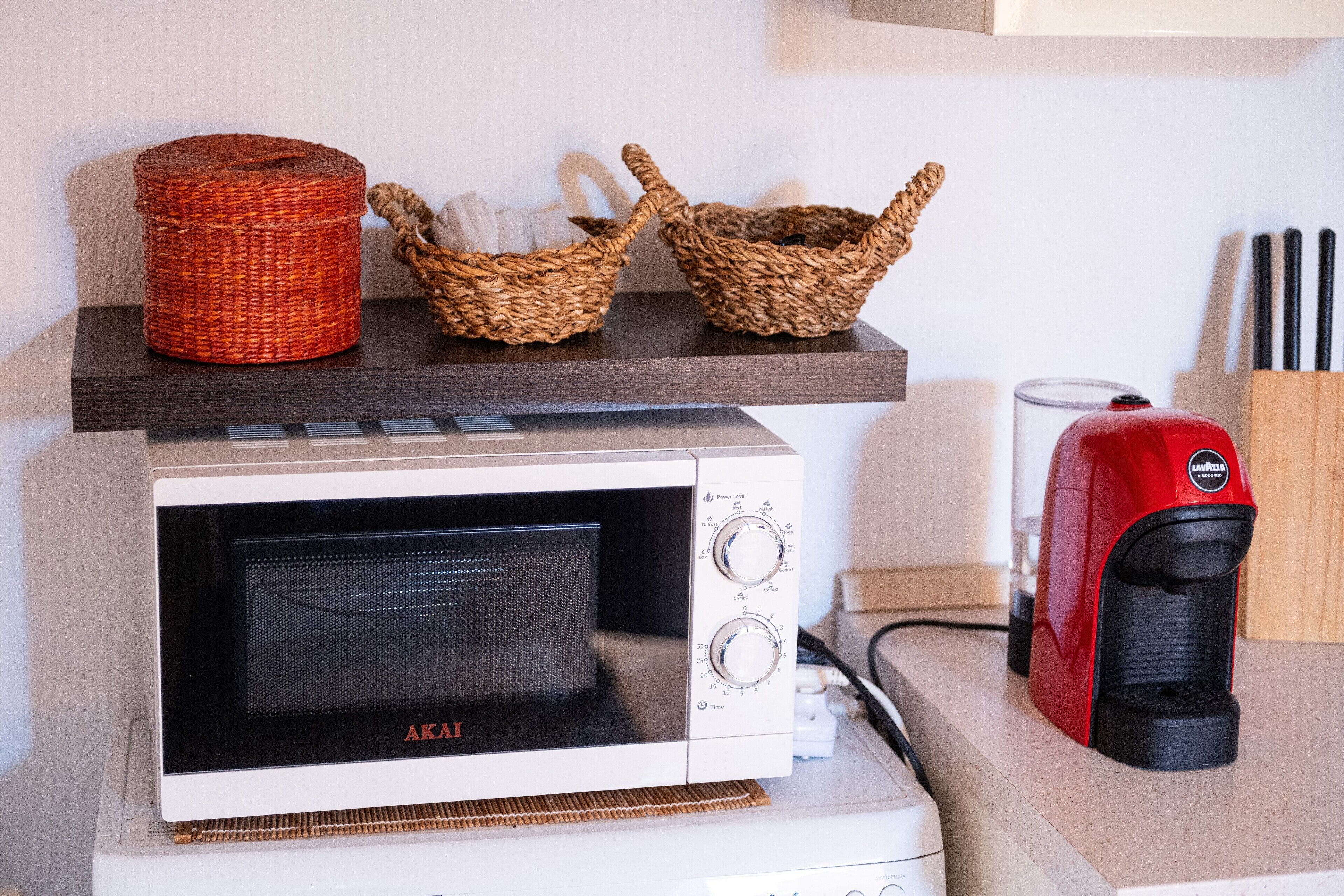 Un horno microondas sobre una encimera con una cafetera roja y dos cestas tejidas en un estante superior.