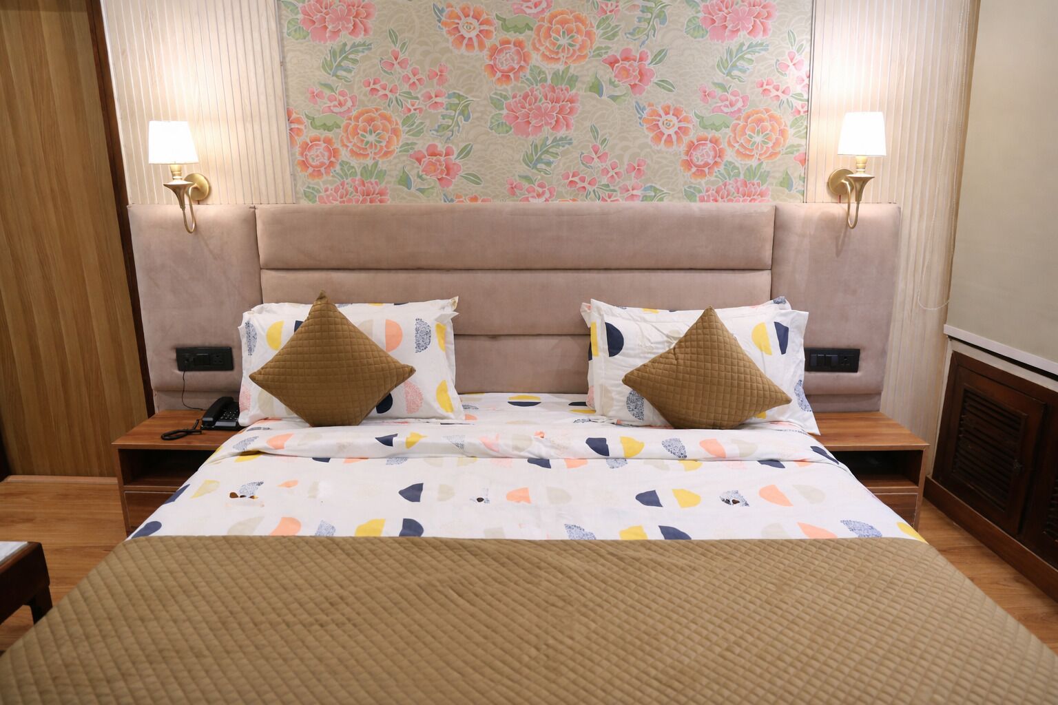 A hotel room with a bed, two bedside lamps, a floral wallpaper, and a wooden nightstand.