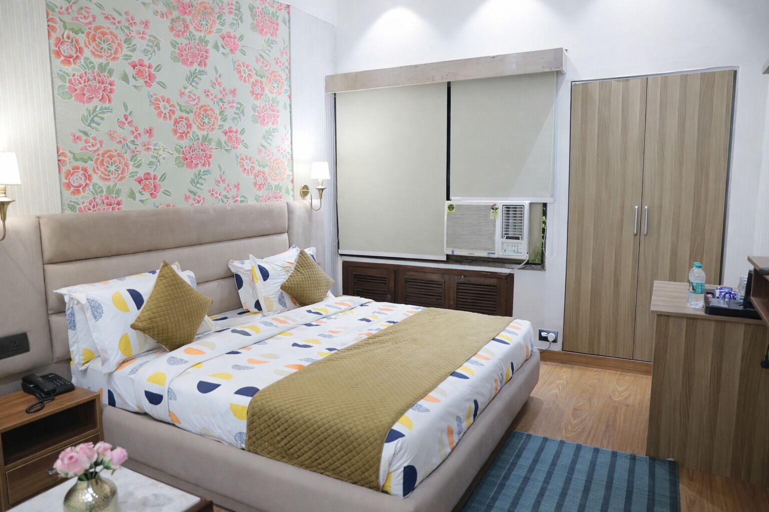 A hotel room with a bed, bedside table, lamp, wardrobe, and an air conditioning unit.