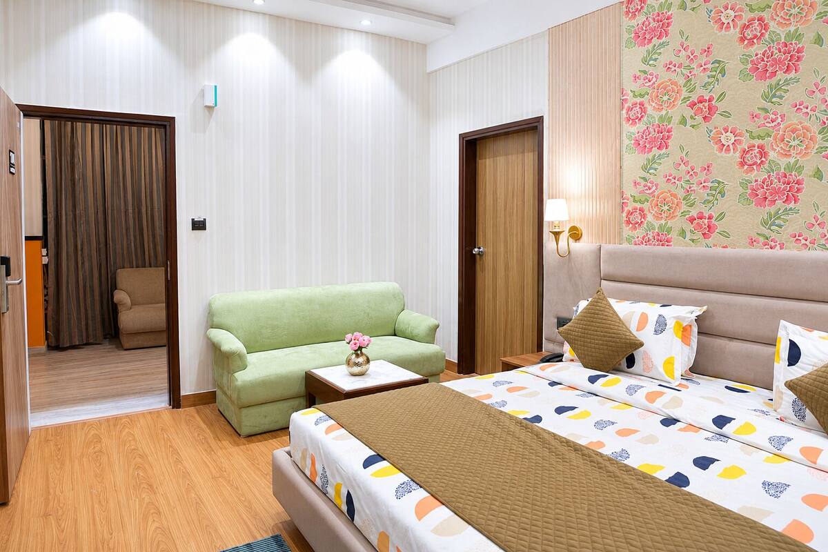 A hotel room with a bed, a green sofa, a small table with a vase, and a wooden wardrobe.
