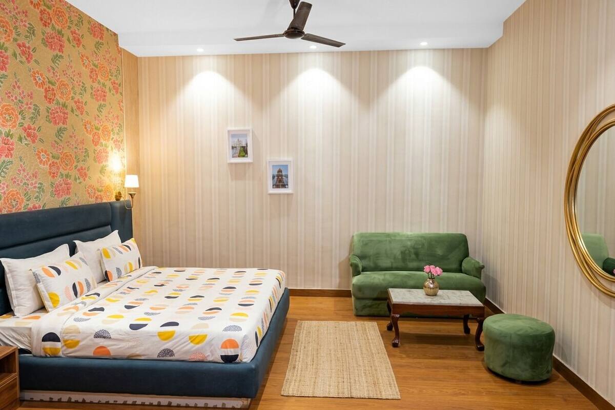 A bedroom with a bed, a green armchair, a small table with a vase, and a ceiling fan.