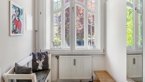 Une pièce avec un radiateur blanc, un banc, une fenêtre donnant sur des arbres et un tableau encadré accroché au mur.