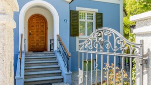 Un bâtiment bleu avec une porte en bois, un escalier doté d’une rampe en métal et une entrée fermée par un portail.