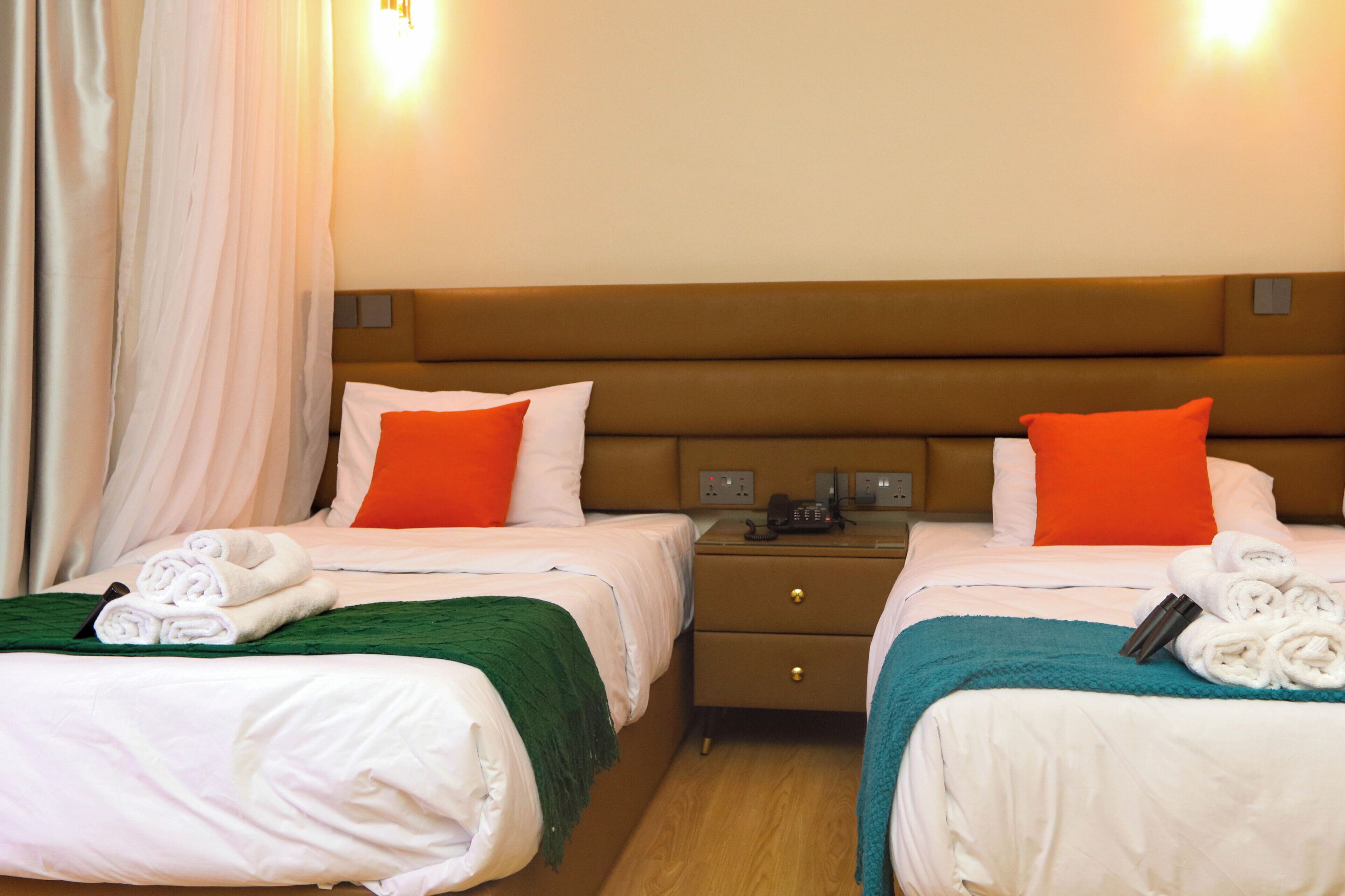A hotel room with two beds, each with a green and white blanket, white pillows, and orange accent pillows. There is a nightstand with a phone and a lamp between the beds.