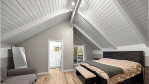 Ein Schlafzimmer mit schrägem Dach, einem Bett mit Holz-Kopfteil, einer Bank, einem grauen Sofa und einem durch eine offene Tür einsehbaren Badezimmer.