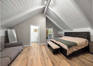 A bedroom with a sloped ceiling, a bed with a wooden headboard, a bench, a gray sofa, and a bathroom visible through an open door.