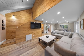 A spacious living area with a high ceiling, wooden paneling, a flat-screen TV, a sofa, a coffee table, and a dining area.
