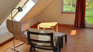 Una habitació amb sostre inclinat, terra de fusta, una cadira, una taula, una làmpada de peu i una finestra amb vista a vegetació.