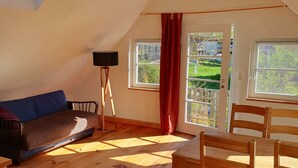 Un habitació d'estil loft amb sostre inclinat, sòl de fusta, un sofà, taula i cadires, i vista a un jardí per una finestra.